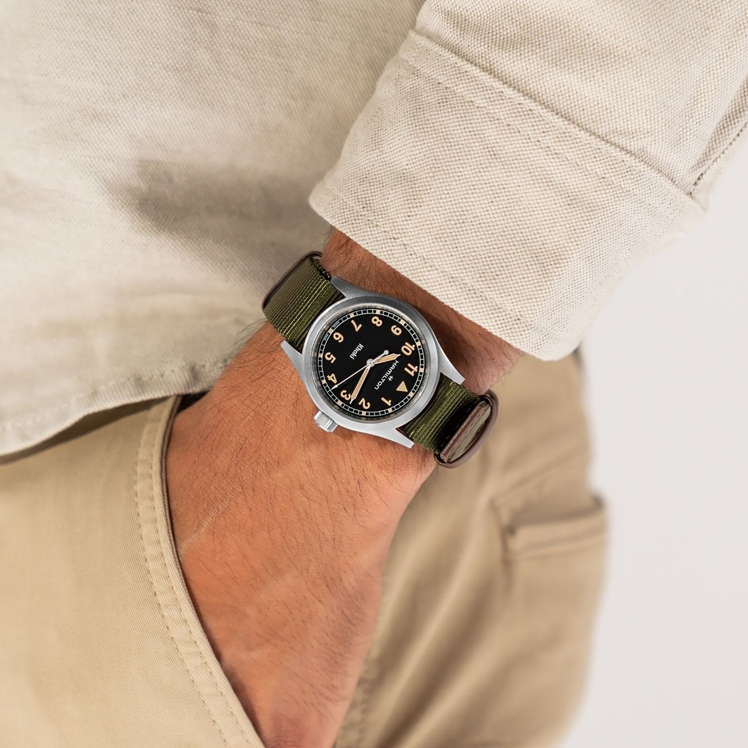 A person wearing a Hamilton Khaki Field Quartz watch with a green strap and black dial, hand in pocket, beige shirt and pants.