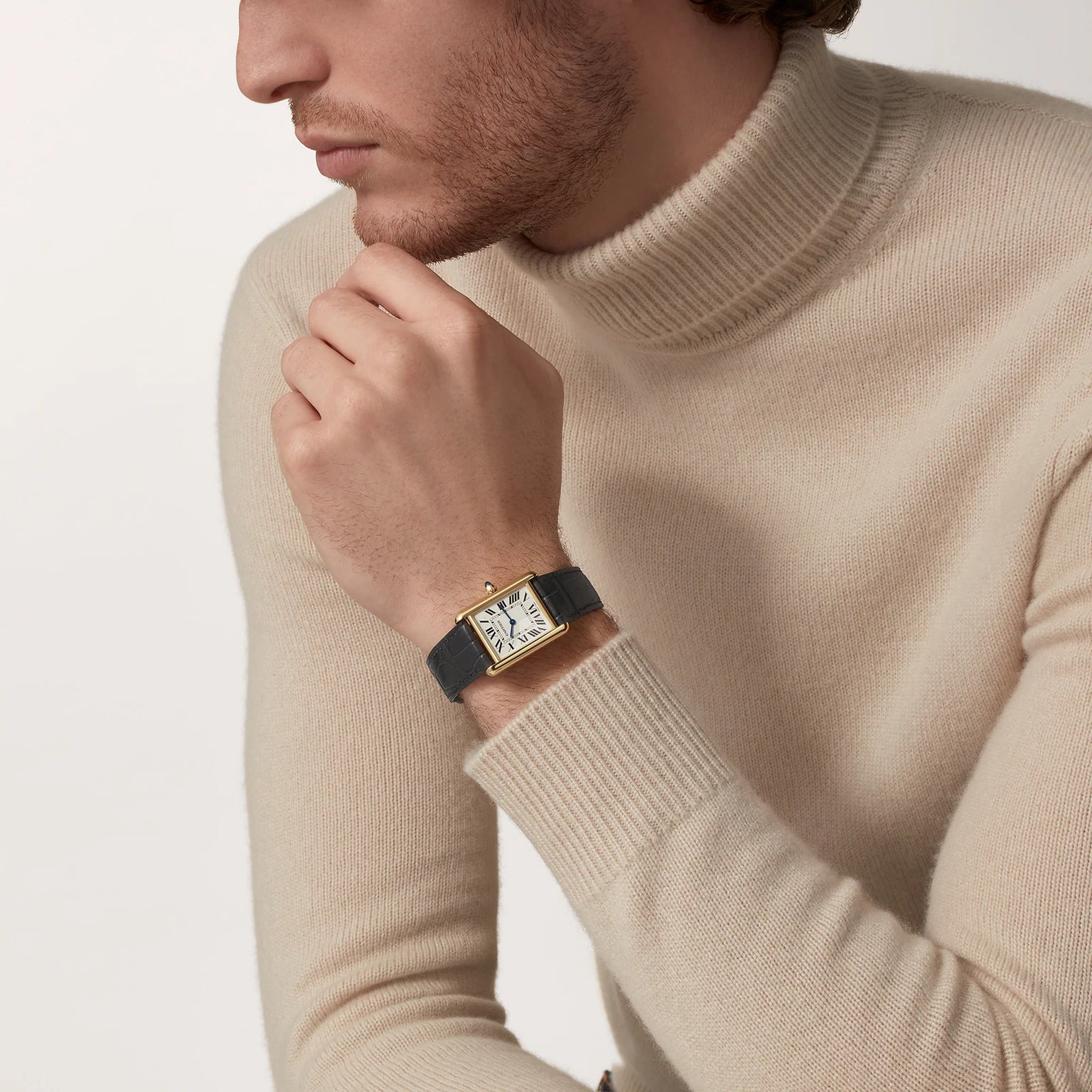 A man in a beige turtleneck poses with his hand near his mouth, showcasing the Cartier Tank Louis Medium Model—a gold watch with a black leather strap that epitomizes modern watchmaking inspired by Louis Cartier.