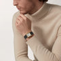 A man in a beige turtleneck poses with his hand near his mouth, showcasing the Cartier Tank Louis Medium Model—a gold watch with a black leather strap that epitomizes modern watchmaking inspired by Louis Cartier.