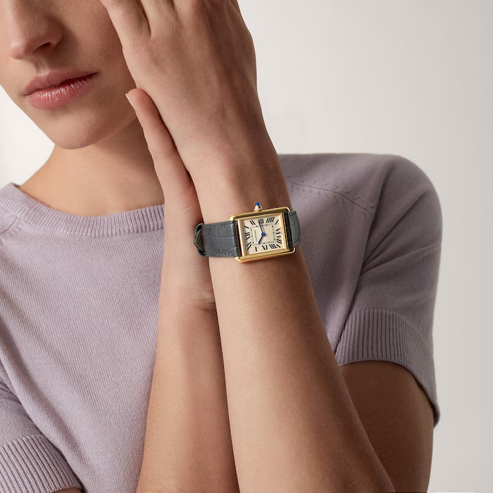A person in a short-sleeved, light purple top showcases the Cartier Tank Louis Medium Model watch with a rectangular face, gray leather strap, and Roman numerals, while holding their opposite wrist.