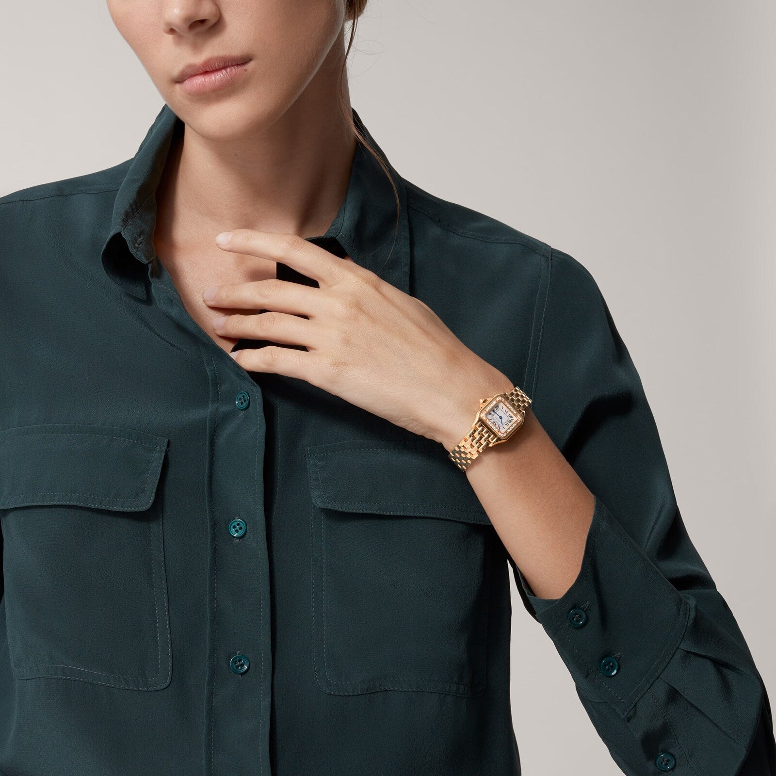 A woman in a dark green button-up shirt showcases a Cartier Panthère de Cartier Small Model gold wristwatch on her left wrist, her hand poised near her collar.