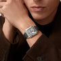 A person in a brown coat wears the Cartier Santos-Dumont Skeleton with Micro-Rotor, featuring a square skeleton dial and black strap on their left wrist, showcasing refined elegance.