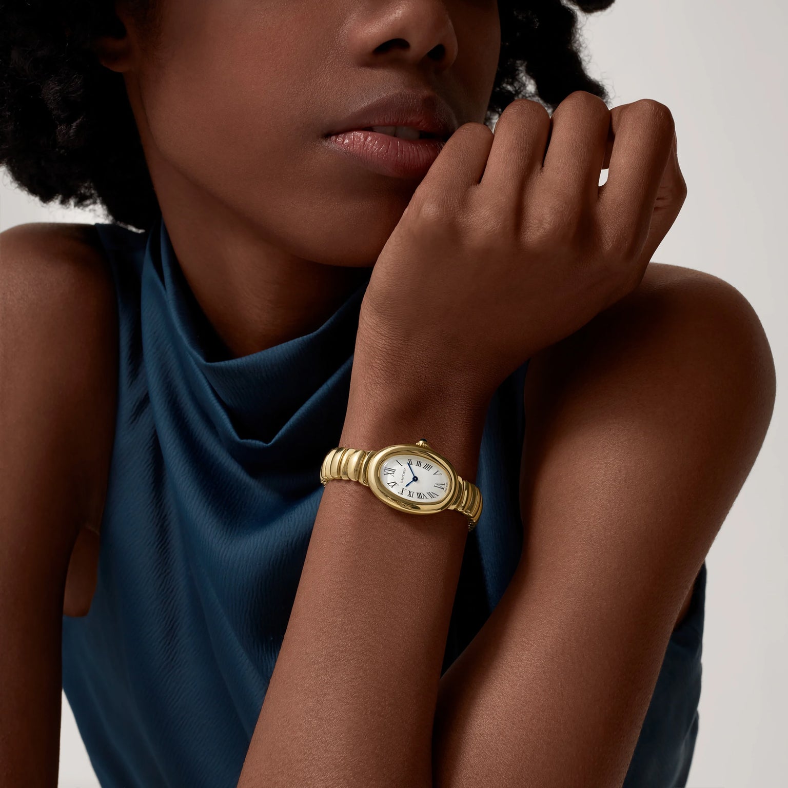 A woman in a sleeveless blue top rests her chin on her hand, wearing a Cartier Baignoire watch in 18K yellow gold with a white analog face and Roman numerals.
