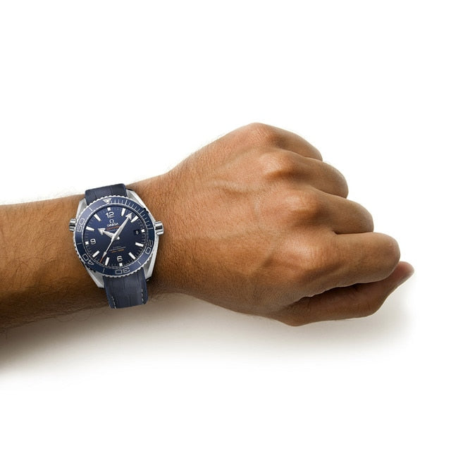 A person’s left wrist wearing an OMEGA Seamaster Planet Ocean 600M watch with a round face and dark blue color, set against a plain white background.