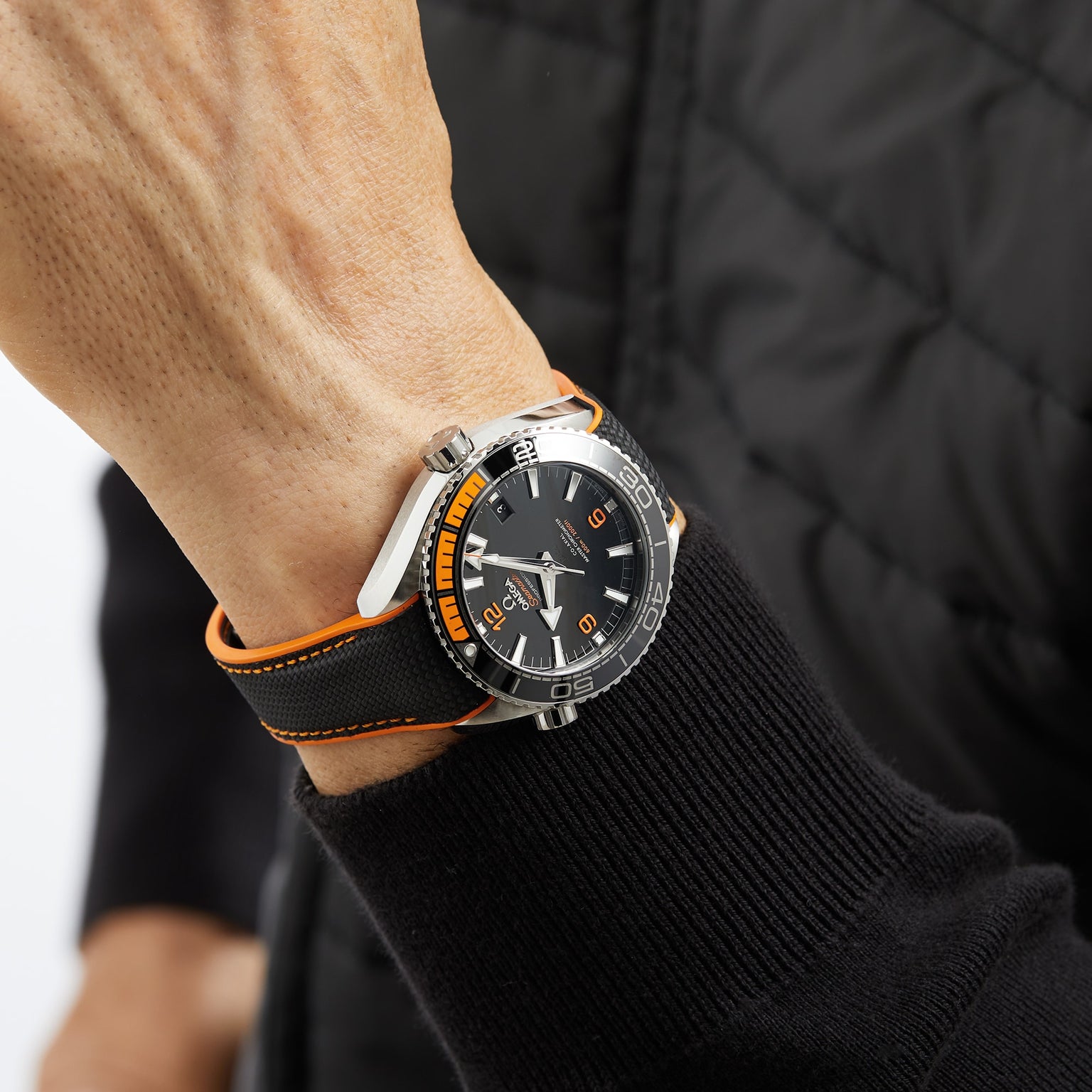 A person’s wrist wearing an OMEGA Seamaster Planet Ocean 600M dive watch with a black and orange bezel, black dial, and a black quilted jacket sleeve visible.