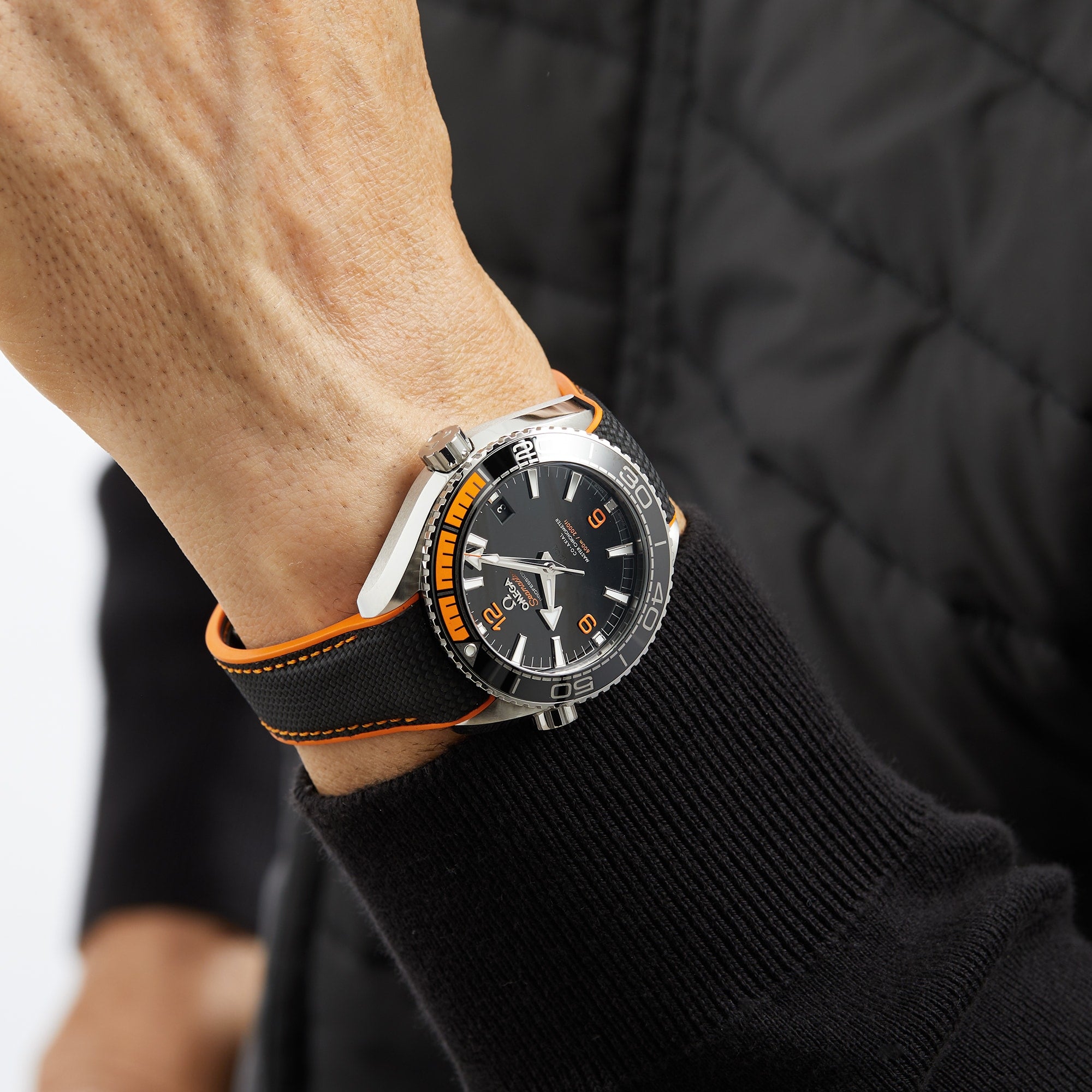 A person’s wrist wearing an OMEGA Seamaster Planet Ocean 600M dive watch with a black and orange bezel, black dial, and a black quilted jacket sleeve visible.