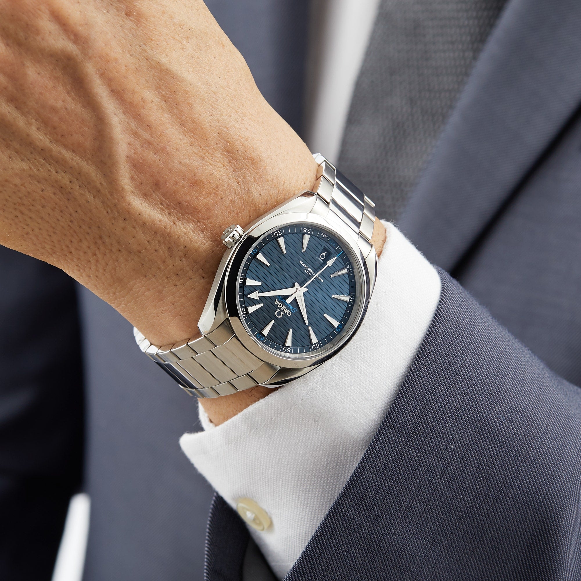 A person dressed in a navy suit and white shirt wears an OMEGA Seamaster Aqua Terra 150M, featuring a stainless steel case and blue patterned dial with silver markers.