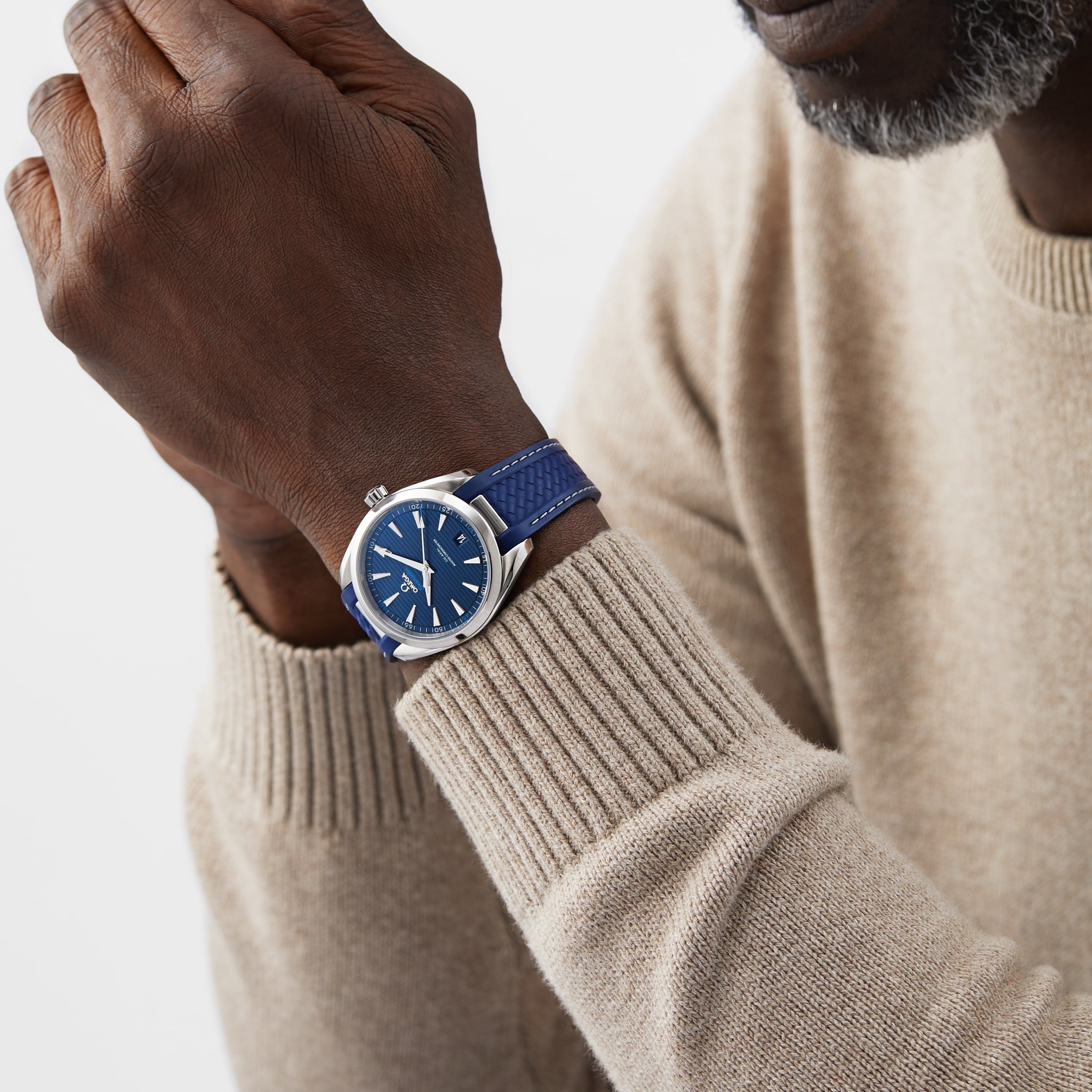 A person in a beige sweater showcases an OMEGA Seamaster Aqua Terra 150M with a blue dial and matching blue strap on their left wrist.