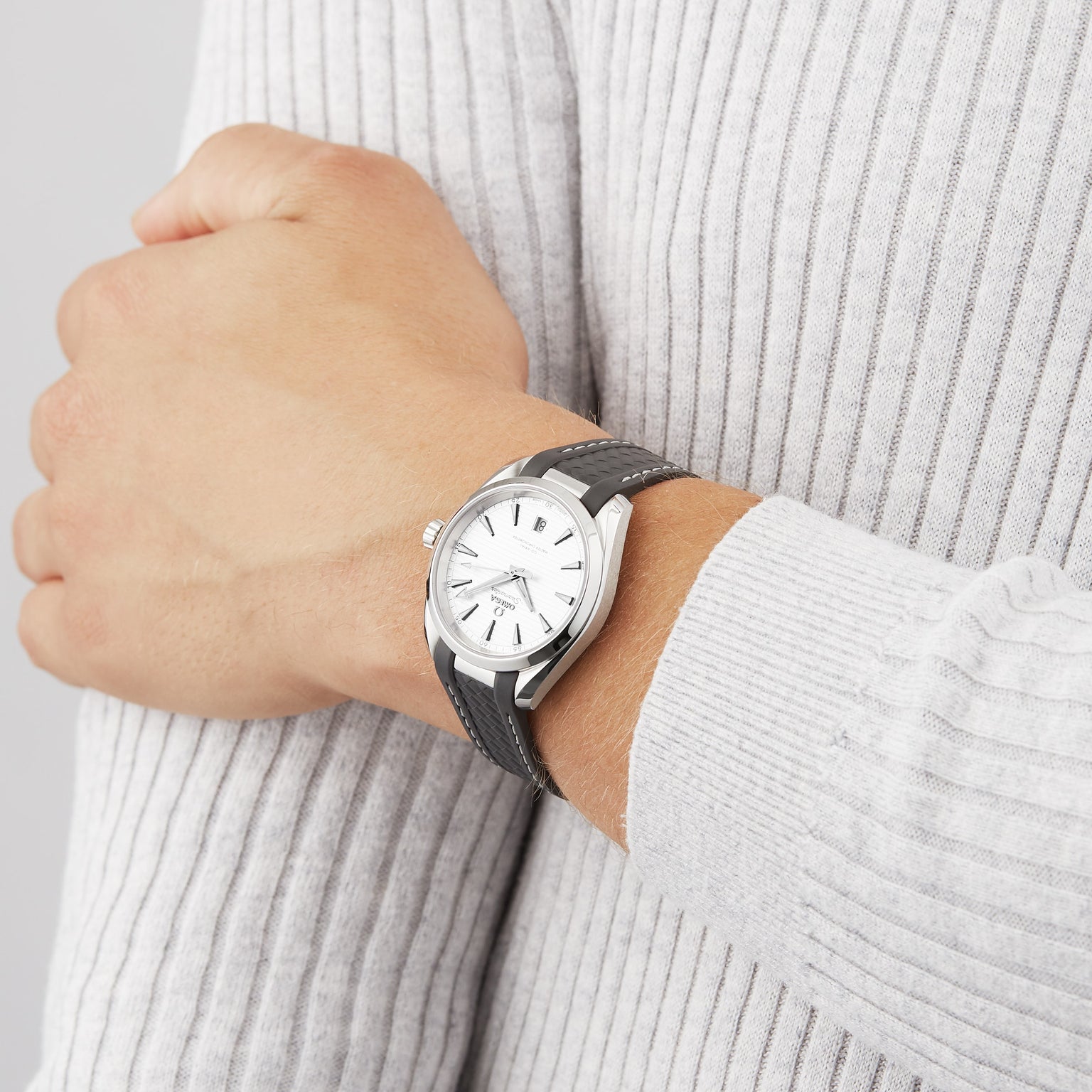 A person wearing an OMEGA Seamaster Aqua Terra 150M wristwatch with a black strap and white dial, dressed in a light gray ribbed sweater.