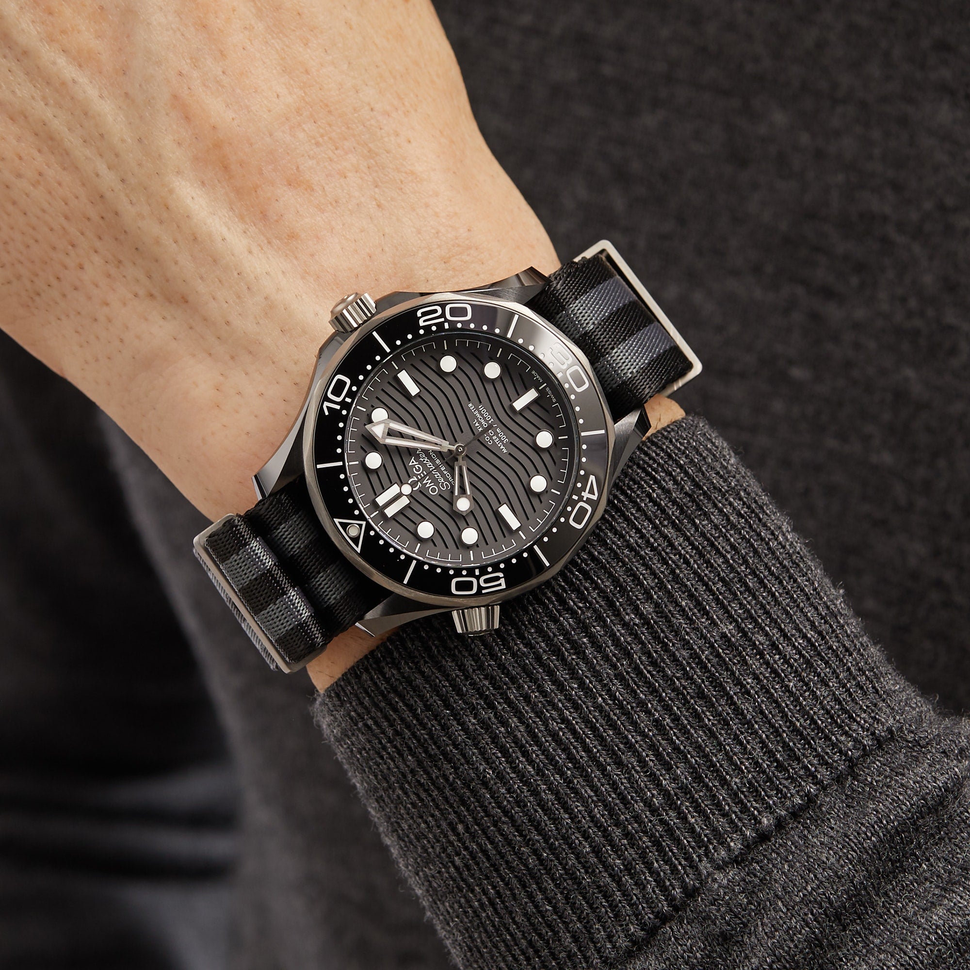 A person wearing an OMEGA Seamaster Diver 300M with a black dial, white markers, stainless steel case, and a black and gray striped strap over a dark long sleeve shirt.
