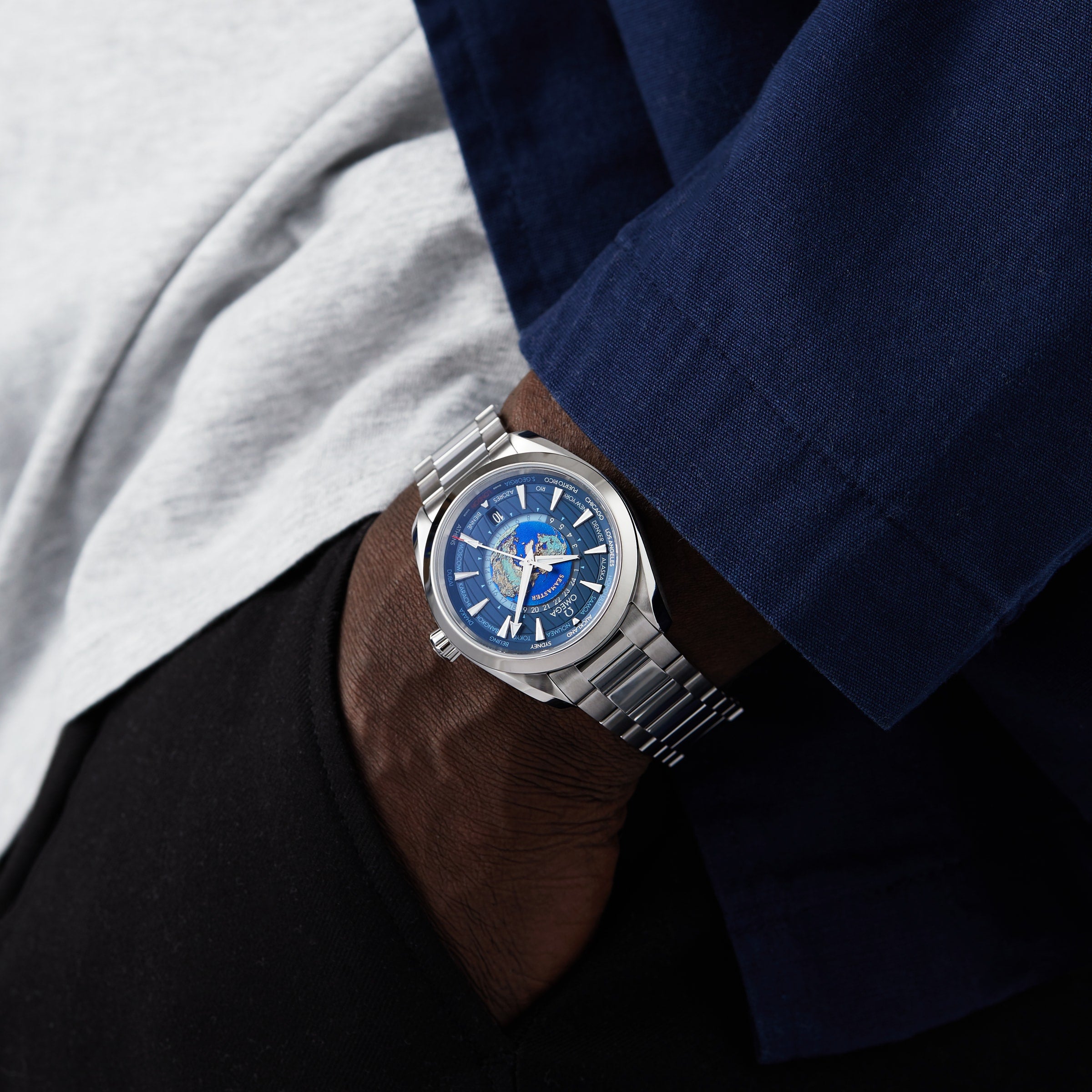 A person wearing an OMEGA Seamaster Aqua Terra 150M with a blue dial and stainless steel band, their hand in the pocket of black pants, dressed in a blue shirt over a gray undershirt.