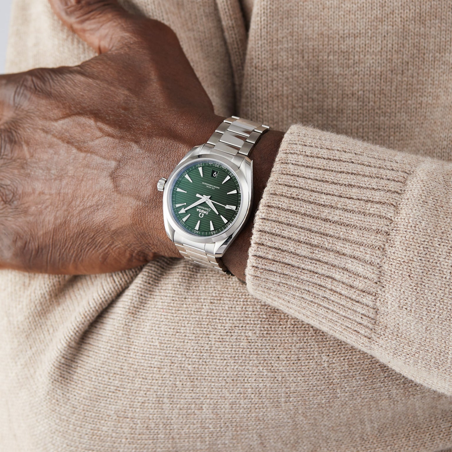 A person in a beige sweater wears an OMEGA Seamaster Aqua Terra 150M with a green dial, featuring the Master Chronometer 8900 calibre, and crosses their arm over their chest.