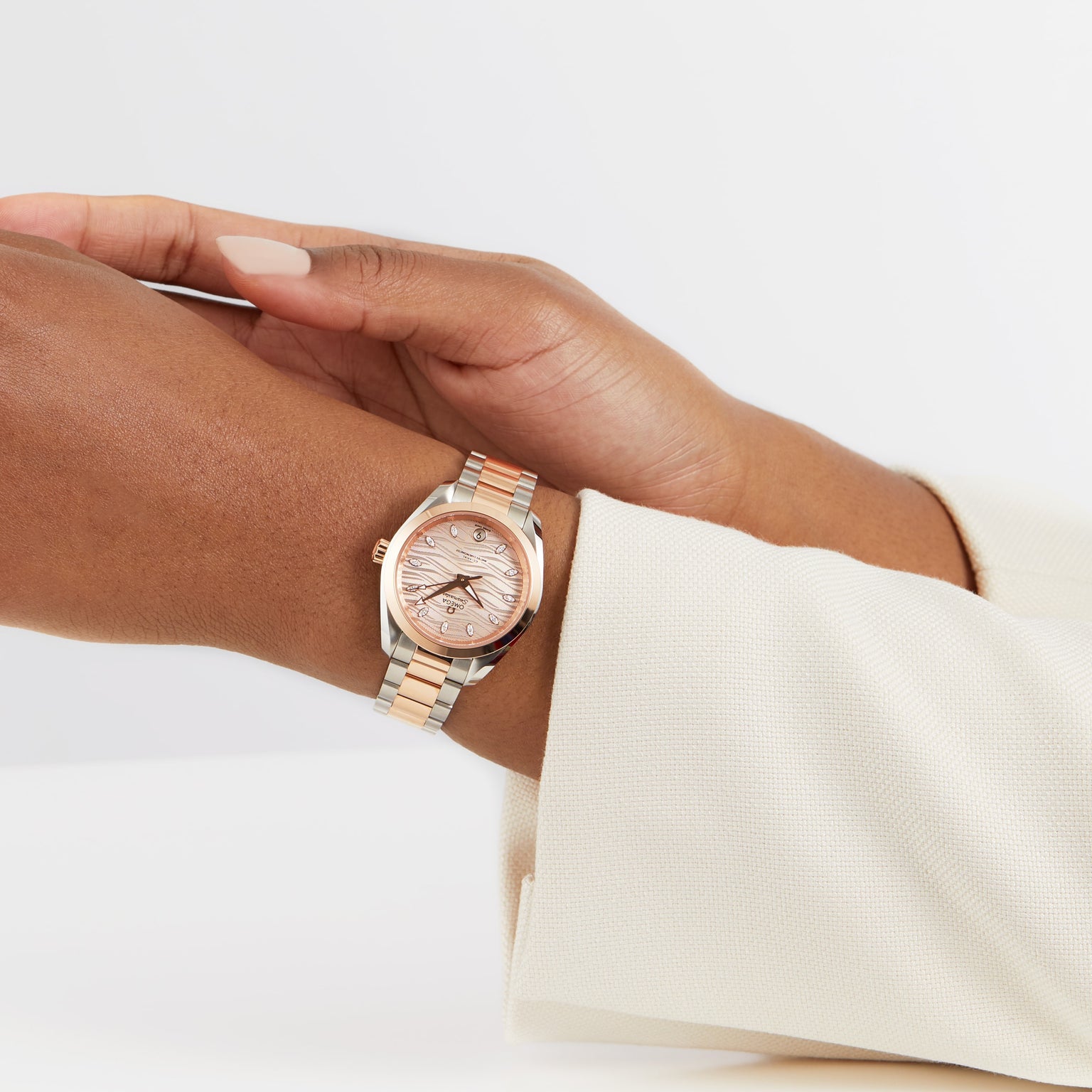A person wearing an OMEGA Seamaster Aqua Terra 150M with a pink dial and gold details adjusts the watch on their left wrist, dressed in a cream-colored long sleeve.