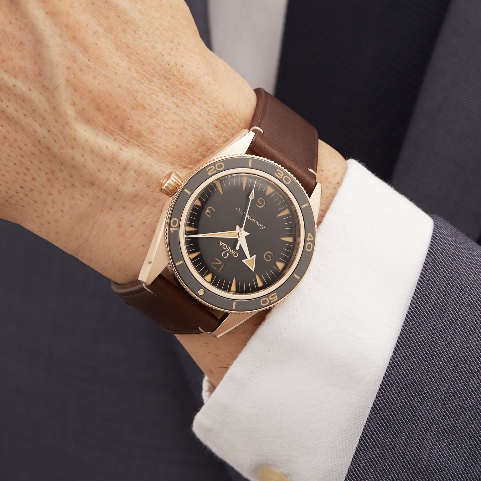 A close-up of a person wearing the OMEGA Seamaster 300 wristwatch with a brown leather strap and bronze gold case, dressed in a white shirt and dark suit jacket.
