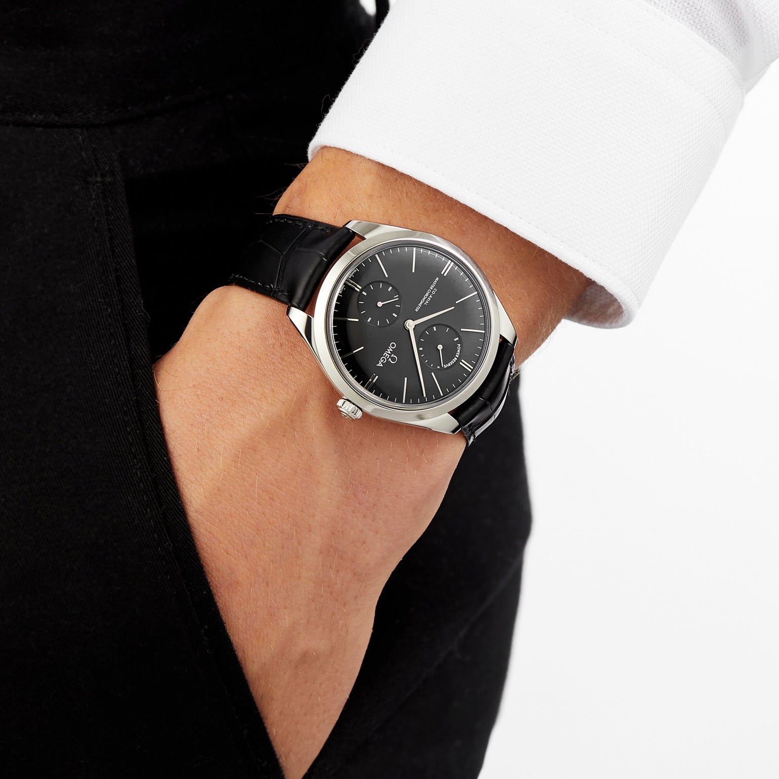 A person in a white long sleeve shirt and black pants has their hand in a pocket, showcasing the OMEGA De Ville Trésor with a 40mm stainless steel case, sleek black dial, and black leather strap.