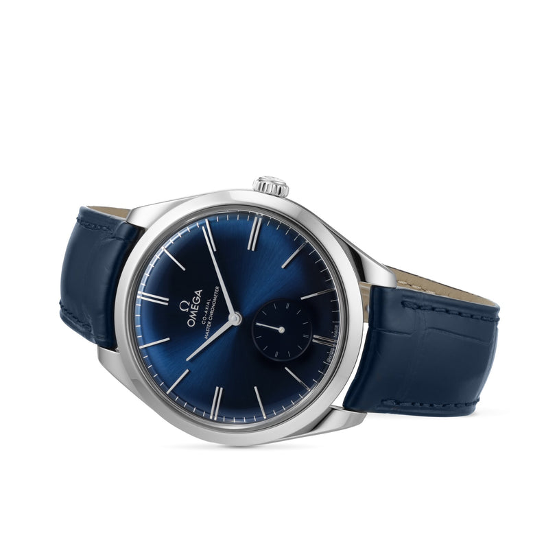 The OMEGA De Ville Trésor, featuring a blue sunburst dial, silver hour markers, and a blue leather strap, rests on its side against a white background. It is powered by the OMEGA Co-Axial Calibre 8926 Master Chronometer movement.