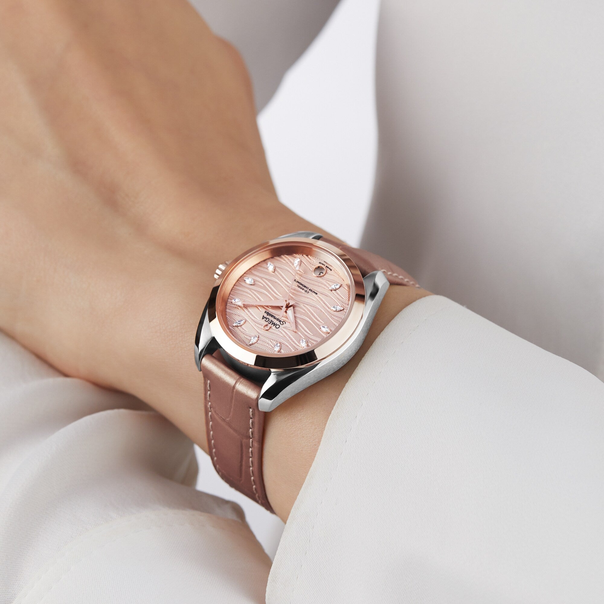 Someone wearing a long-sleeved white shirt and a pink leather OMEGA Seamaster Aqua Terra 150M featuring a rose gold face with a silver case.