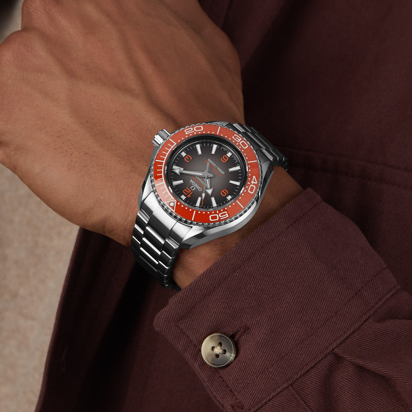 A person in a maroon shirt showcases the OMEGA Seamaster Planet Ocean 6000M—an Ultra Deep stainless steel wristwatch with a black dial, orange bezel, and O-MEGASTEEL construction designed for saturation divers.