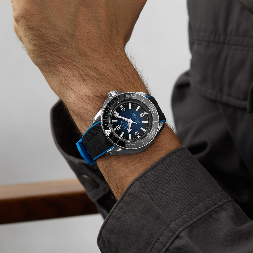 A person wearing an OMEGA Seamaster Planet Ocean 600M with black and silver design, blue accents, and a black strap on their wrist, paired with a dark long-sleeve shirt.