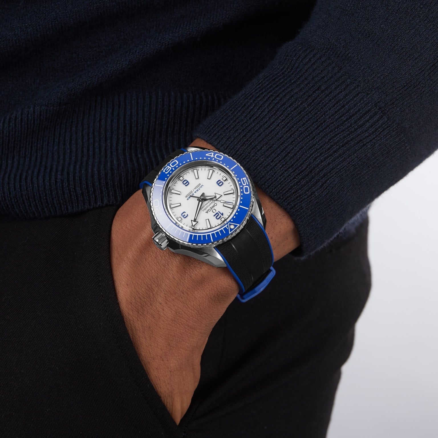 Someone wears an OMEGA Seamaster Planet Ocean 6000M wristwatch with a blue and white face and black band, famed for its water resistance, as their hand rests in the pocket of dark pants.