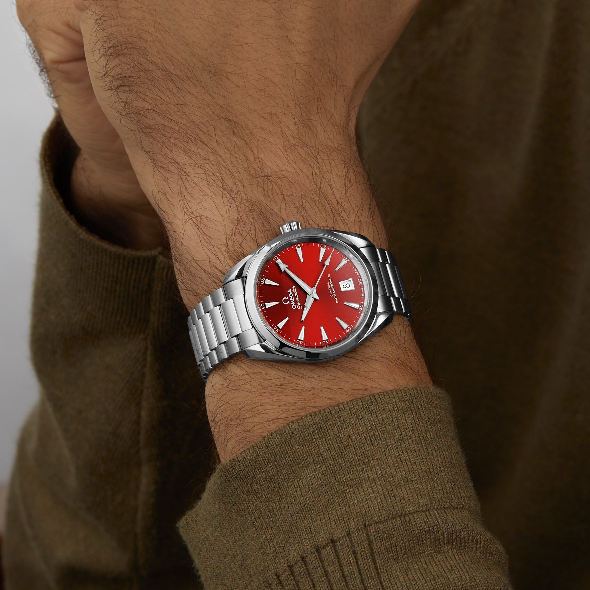 Someone wearing an OMEGA Seamaster Aqua Terra 150M with a silver case, red dial, date display, and precise 8800 calibre movement, paired with a greenish-brown long sleeve shirt.