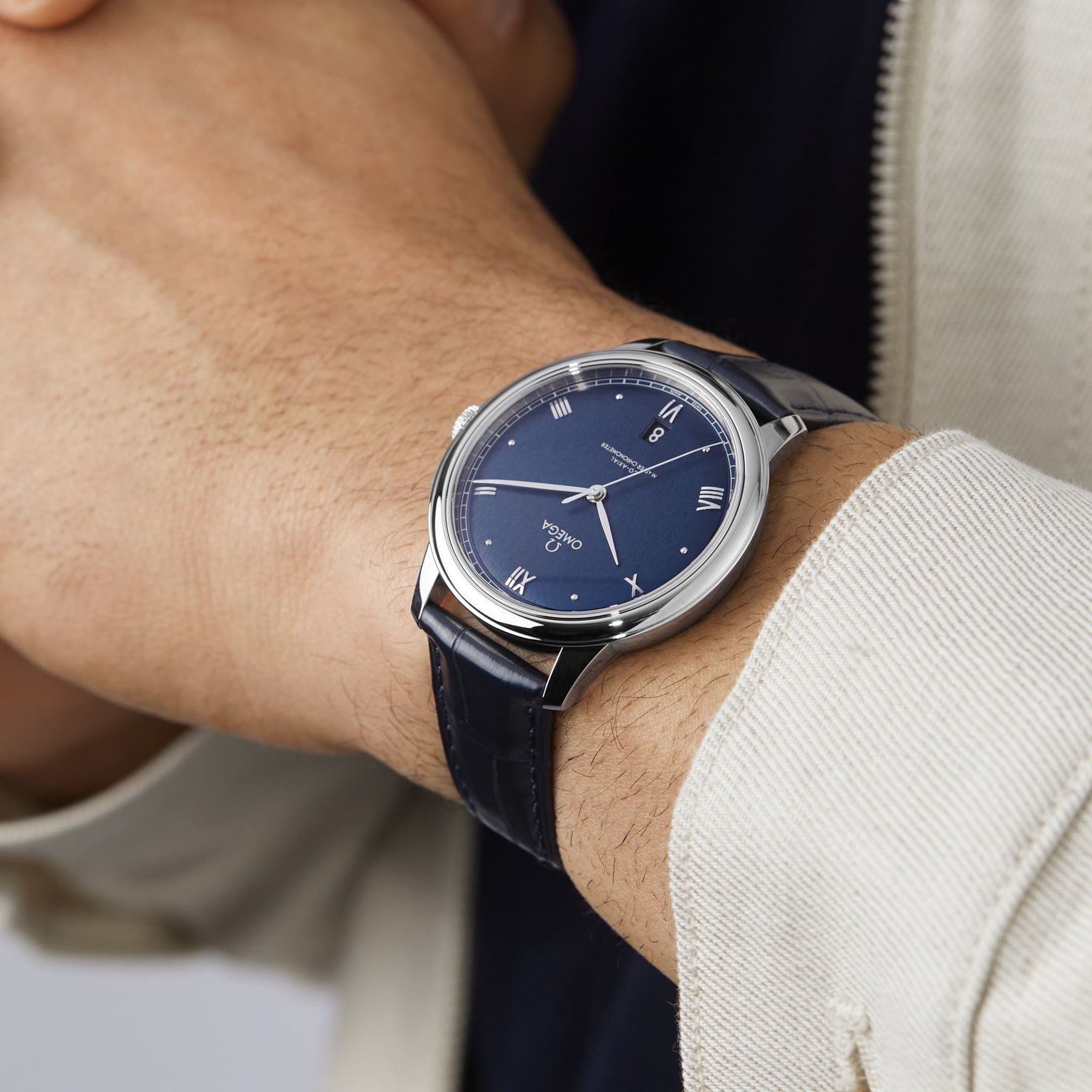 Someone wearing an OMEGA De Ville Prestige with a blue dial, Roman numerals, silver case, and dark blue leather strap—powered by the Co-Axial 8800 calibre—is dressed in a light jacket.