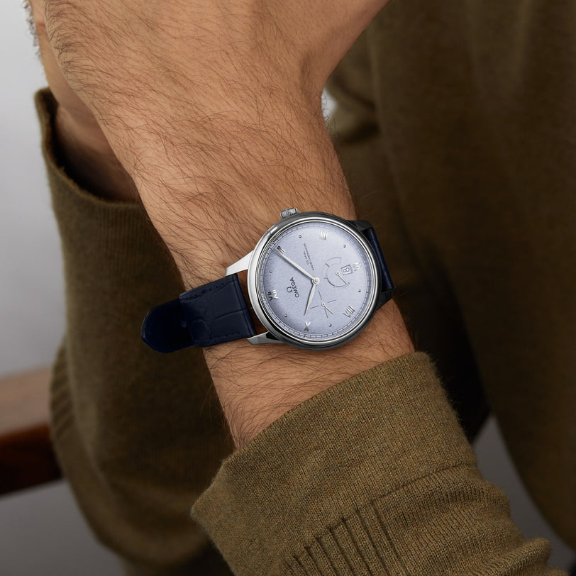 A person in a brown long-sleeve shirt wears an OMEGA De Ville Prestige watch with a stainless steel case, dark leather strap, and light dial.