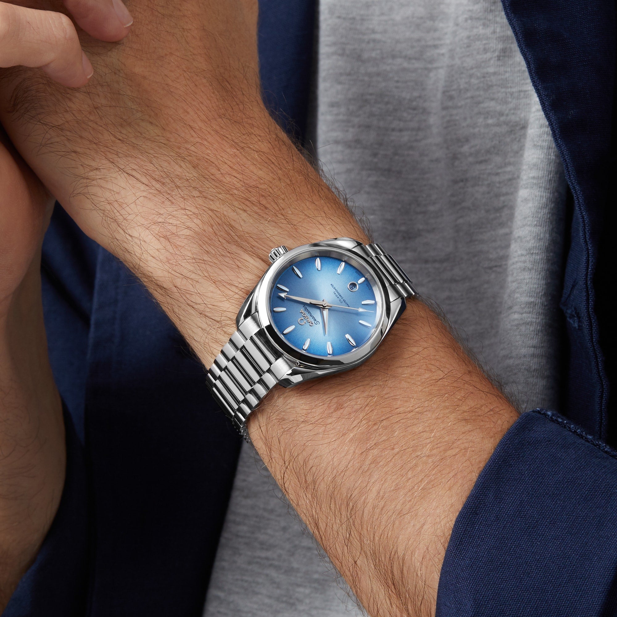A person in a gray shirt and blue jacket wears an OMEGA Seamaster Aqua Terra 150M with a metallic band and blue dial, reflecting maritime heritage.