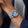 A person in a gray shirt and blue jacket wears an OMEGA Seamaster Aqua Terra 150M with a metallic band and blue dial, reflecting maritime heritage.