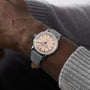 A person wearing a Raymond Weil Millesime Automatic Salmon Sector Dial Swiss-made silver wristwatch with a grey leather strap, dressed in a grey sweater.
