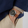 Wearing a Longines Record Chronograph with a brown leather strap, black dial, and stainless steel case, the person pairs it with a blue jacket, beige sweater, and light khaki pants, hand casually in their pocket.