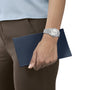 A person wearing a Tissot PRX Powermatic 80 holds a dark blue wallet or clutch against their brown pants and light blue top.