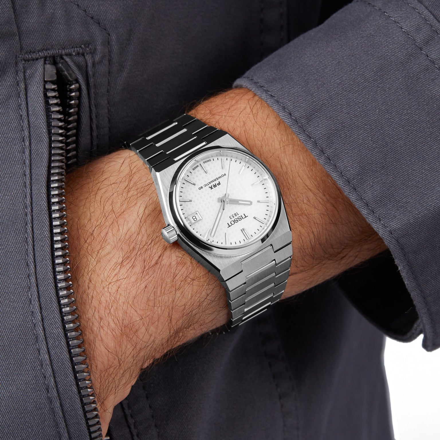 A person in a grey jacket wears a Tissot PRX Powermatic 80 with a 35mm white dial, black hour markers, and date display.