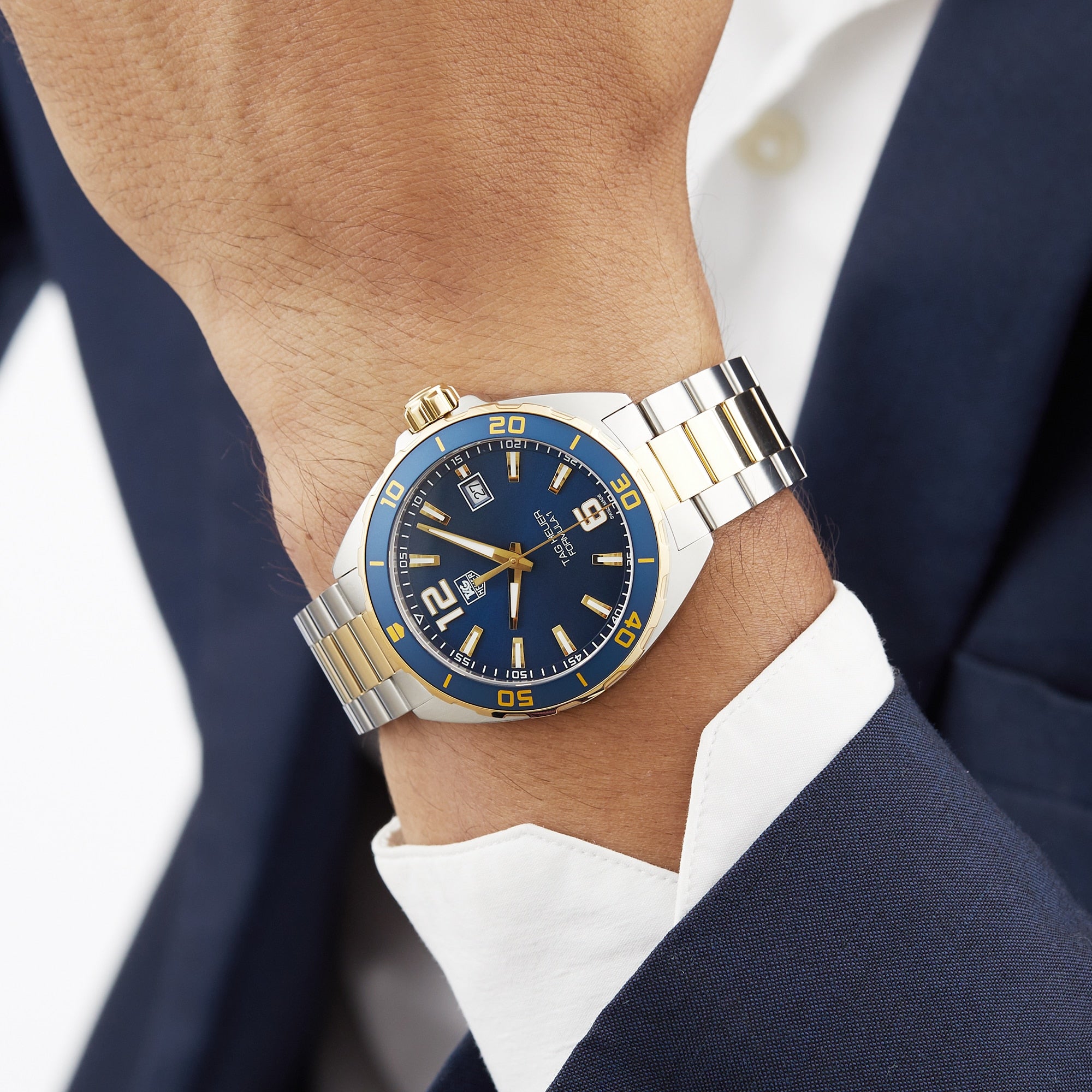 A person wearing the TAG Heuer Special Edition Formula 1 Date, featuring a blue and gold bezel with a blue dial, paired with a navy suit and white shirt.