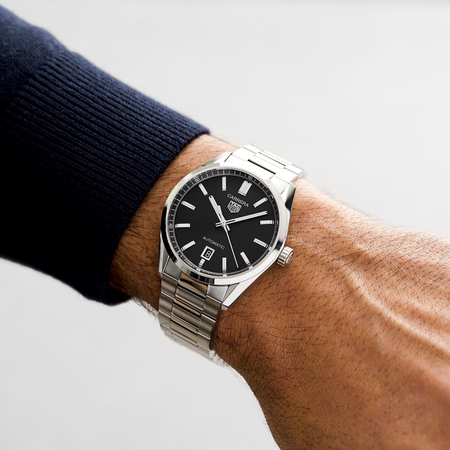 A person wearing a silver TAG Heuer Carrera Date watch with a black dial and date display on their left wrist.