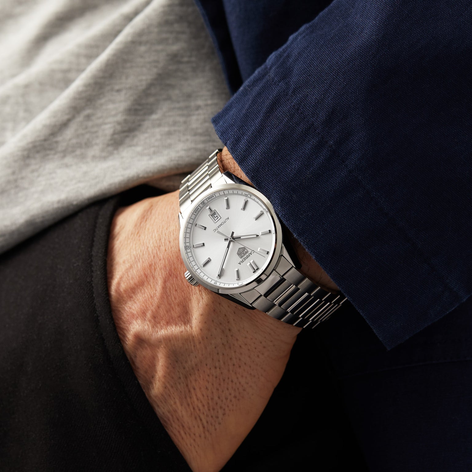 Wearing a TAG Heuer Carrera Date silver wristwatch with a white dial and metal band, the person has their hand in the pocket of dark pants, dressed in a grey shirt and navy jacket.