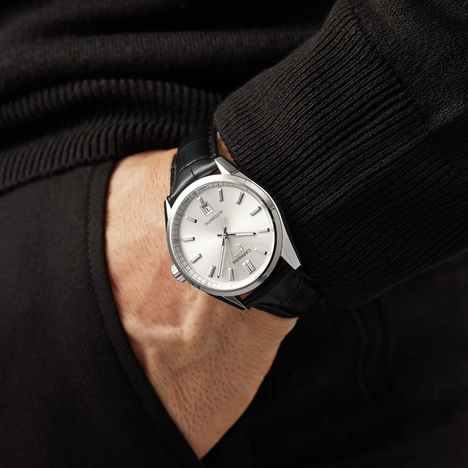 A person in a black sweater and pants, hand in pocket, wears a TAG Heuer Carrera Date analog wristwatch with automatic movement, 39mm steel case, and black leather strap.