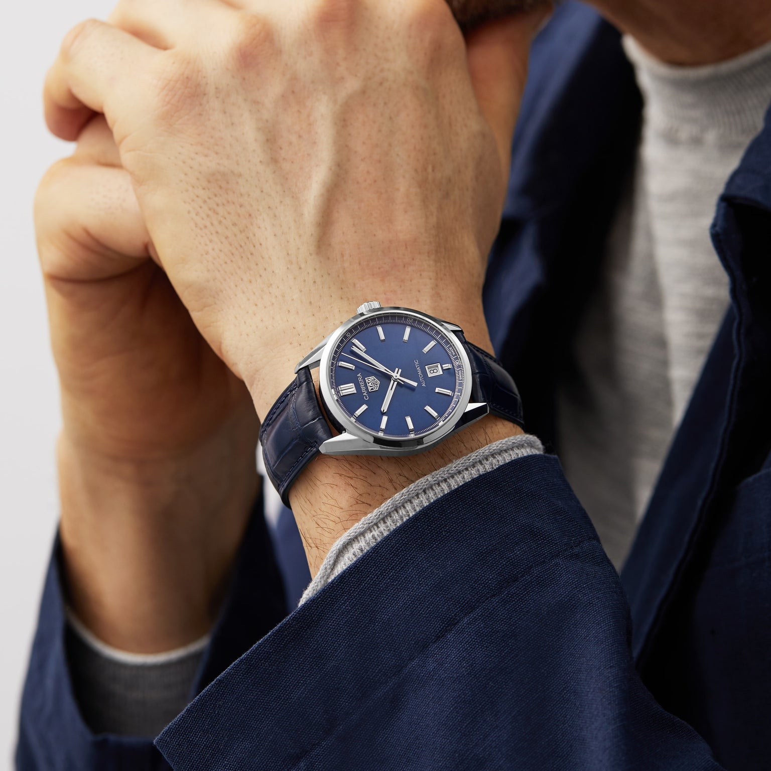 Wearing a TAG Heuer Carrera Date with a blue face and blue leather strap, powered by the calibre 5 automatic movement, a person in a blue jacket and grey shirt rests their chin on their hands—embodying endurance and performance.