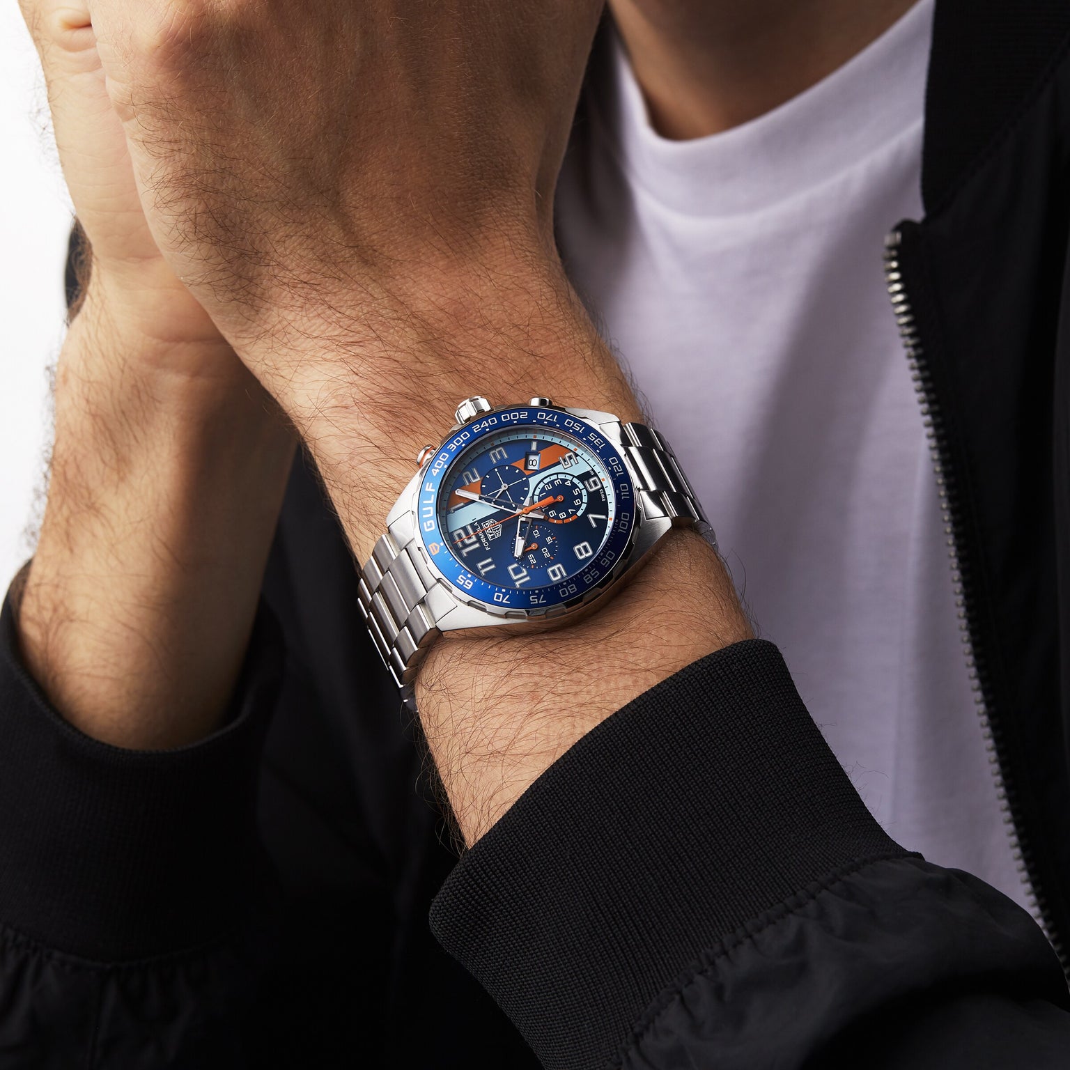 A person in a white shirt and black jacket wears a TAG Heuer Special Edition Formula 1 Chronograph X Gulf wristwatch, featuring a blue face and multiple dials.