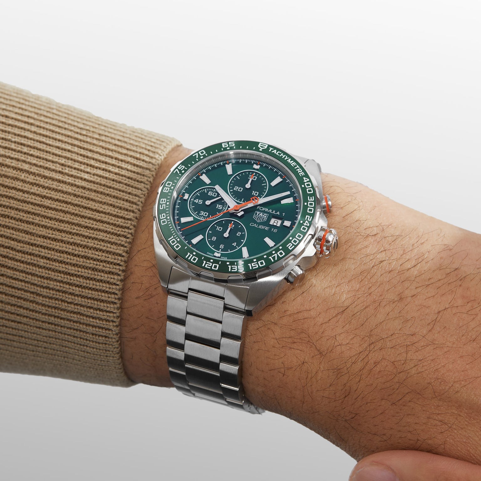 A person in a beige sweater wears the TAG Heuer Formula 1 Chronograph—a silver metal wristwatch with a green ceramic tachymeter bezel, three subdials, and a striking green face.