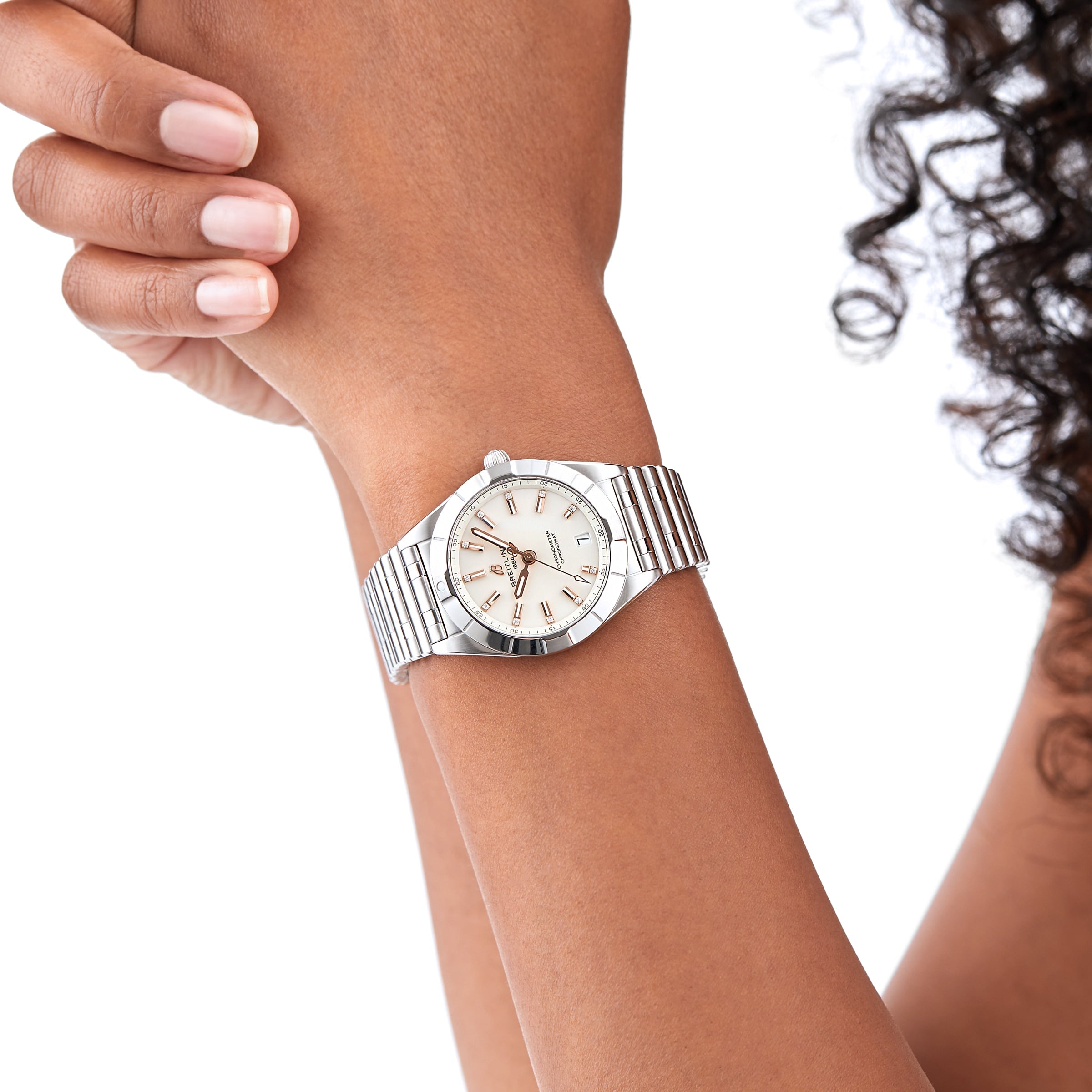 A person with curly hair wears a Breitling Chronomat 32 wristwatch with a silver Rouleaux bracelet, white dial, and Roman numerals, displaying it against a plain white background.