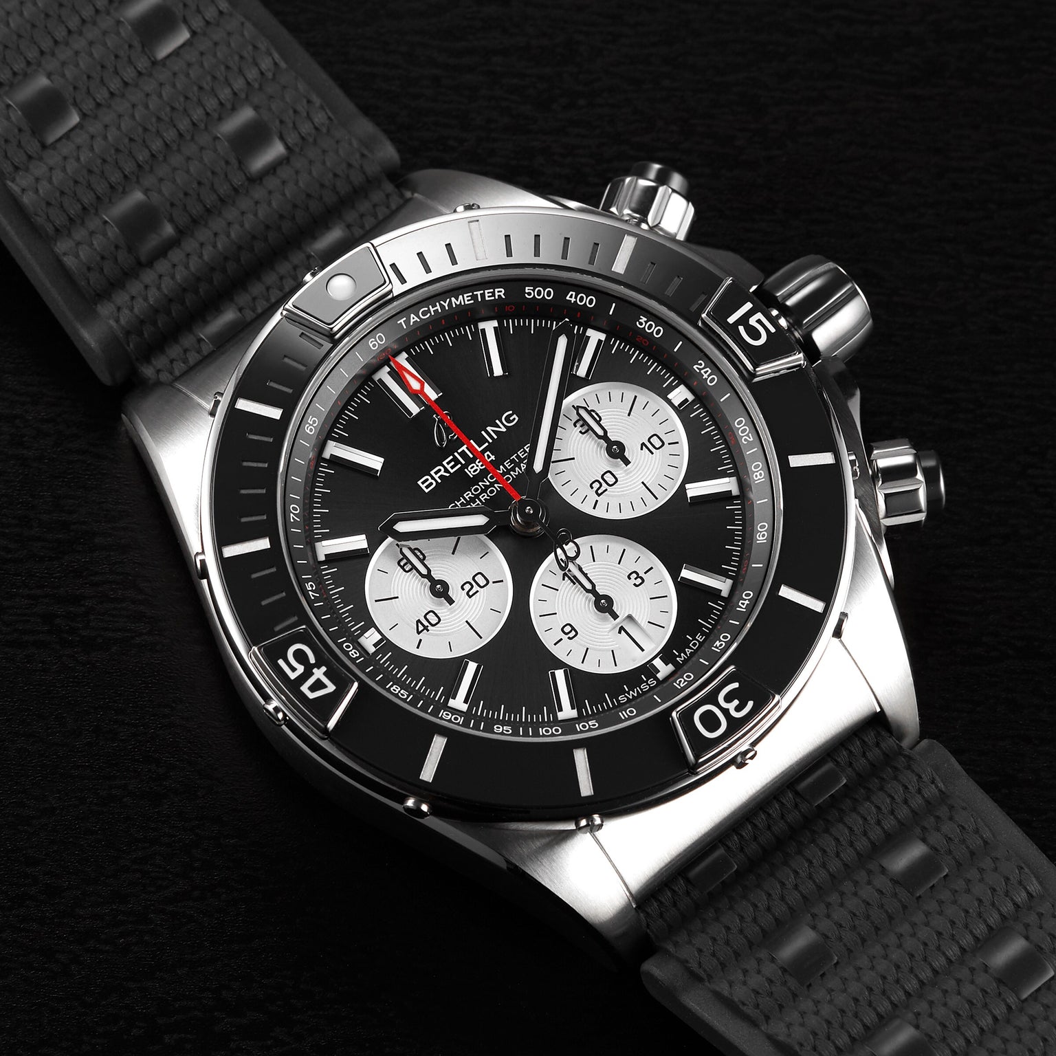 The Breitling Super Chronomat B01 44 chronograph features a black bezel, three sub-dials, a tachymeter scale, and a black textured strap with the precise Breitling 01 COSC-certified calibre, elegantly displayed on a dark surface.
