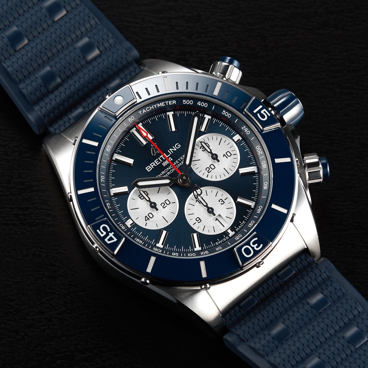 The Breitling Super Chronomat B01 44 is a stainless steel mechanical chronograph wristwatch featuring a blue bezel, black dial with three subdials, and a blue rubber strap.
