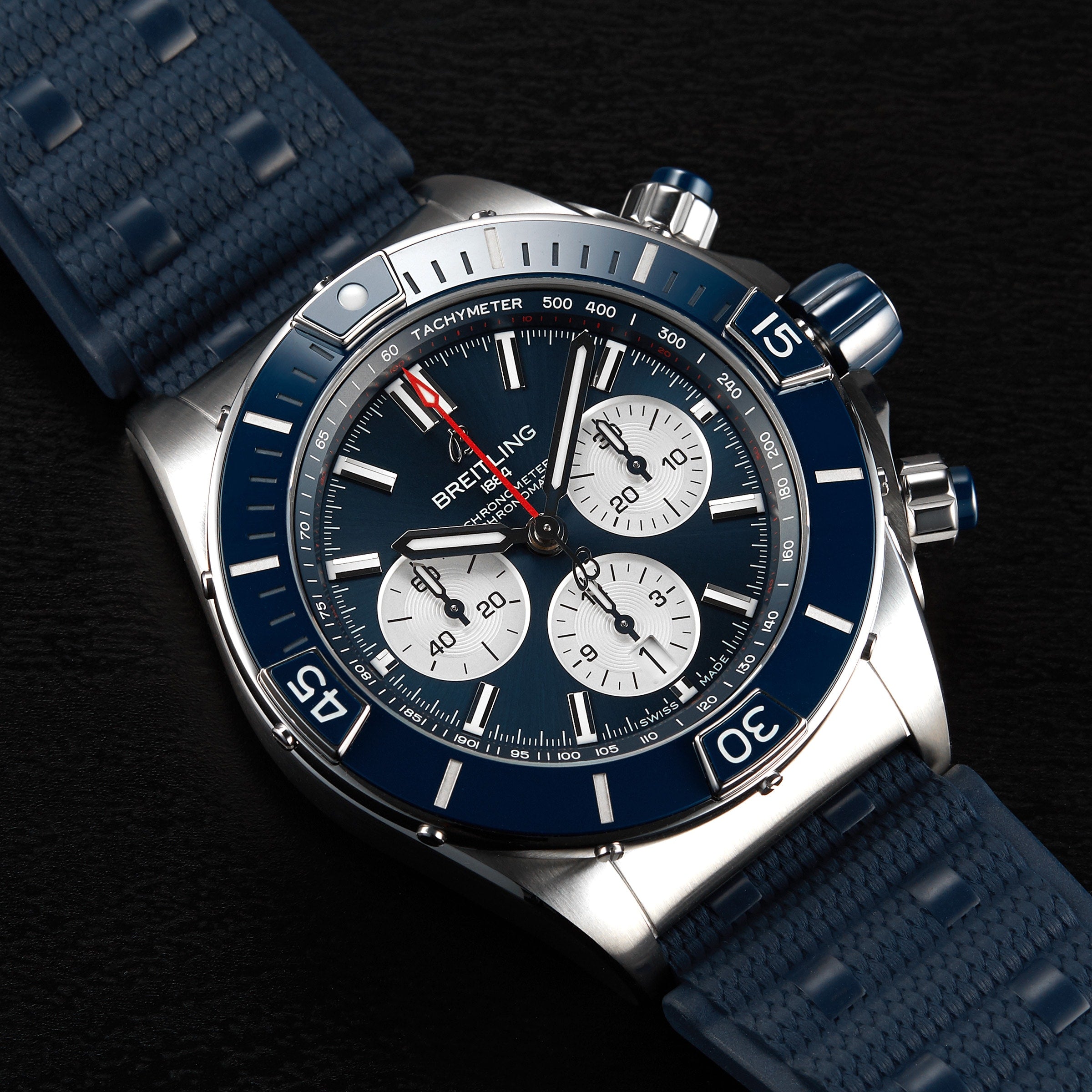 The Breitling Super Chronomat B01 44 is a stainless steel mechanical chronograph wristwatch featuring a blue bezel, black dial with three subdials, and a blue rubber strap.