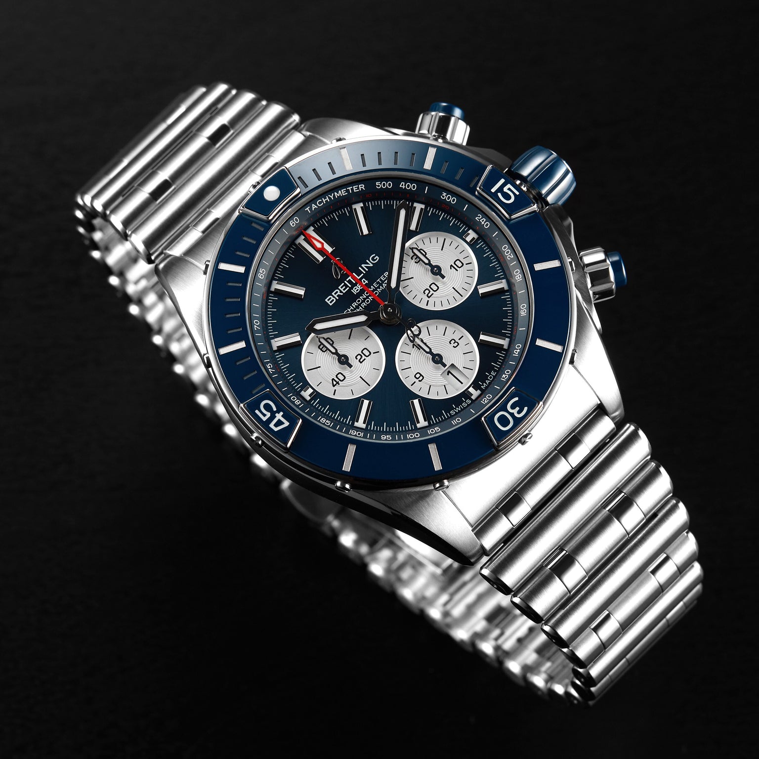 A Breitling Super Chronomat B01 44 stainless steel chronograph wristwatch with a blue bezel, three subdials, and a metal bracelet is displayed on a black surface.
