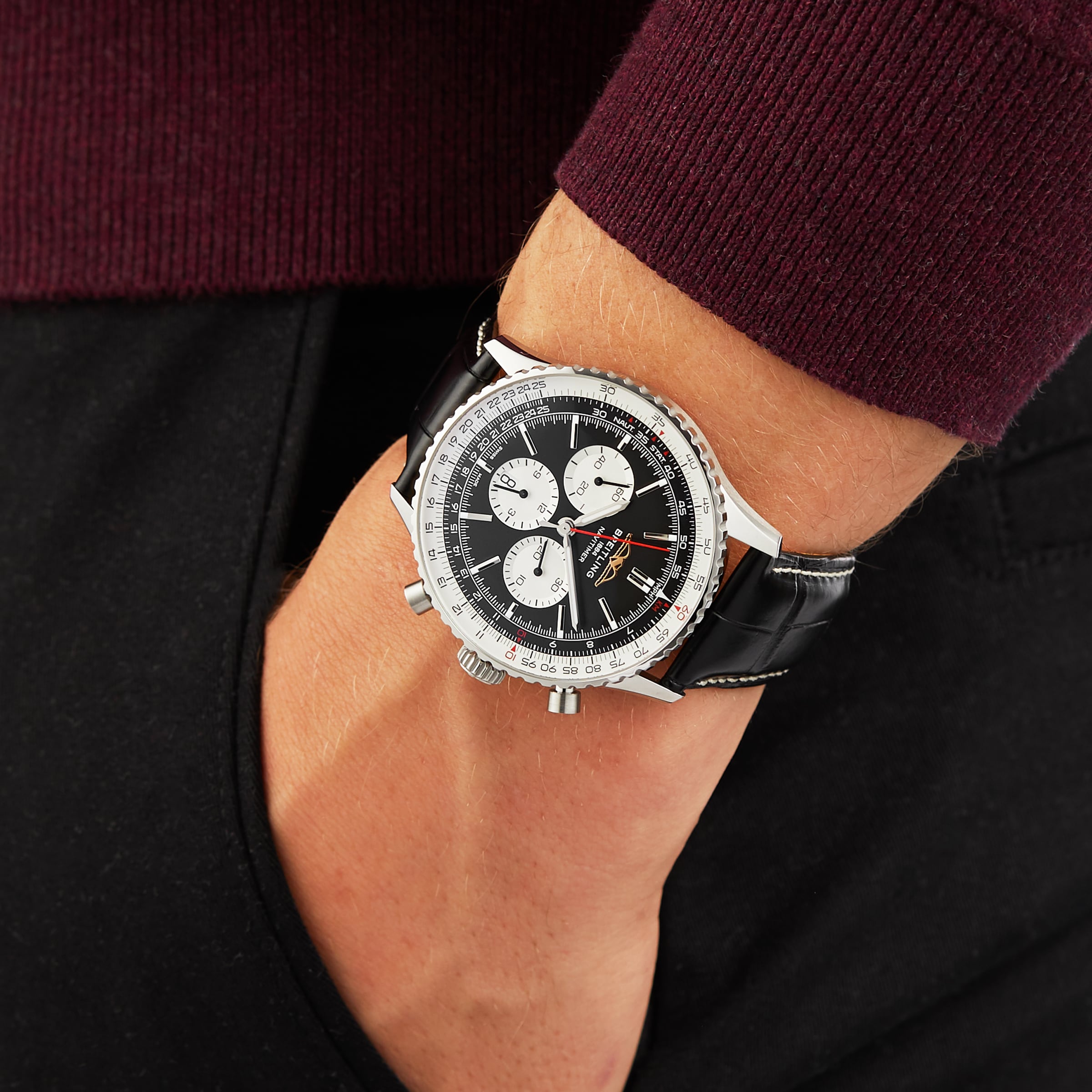A person wearing a Breitling Navitimer B01 Chronograph 46 with a black leather strap, paired with a maroon sweater and black pants.