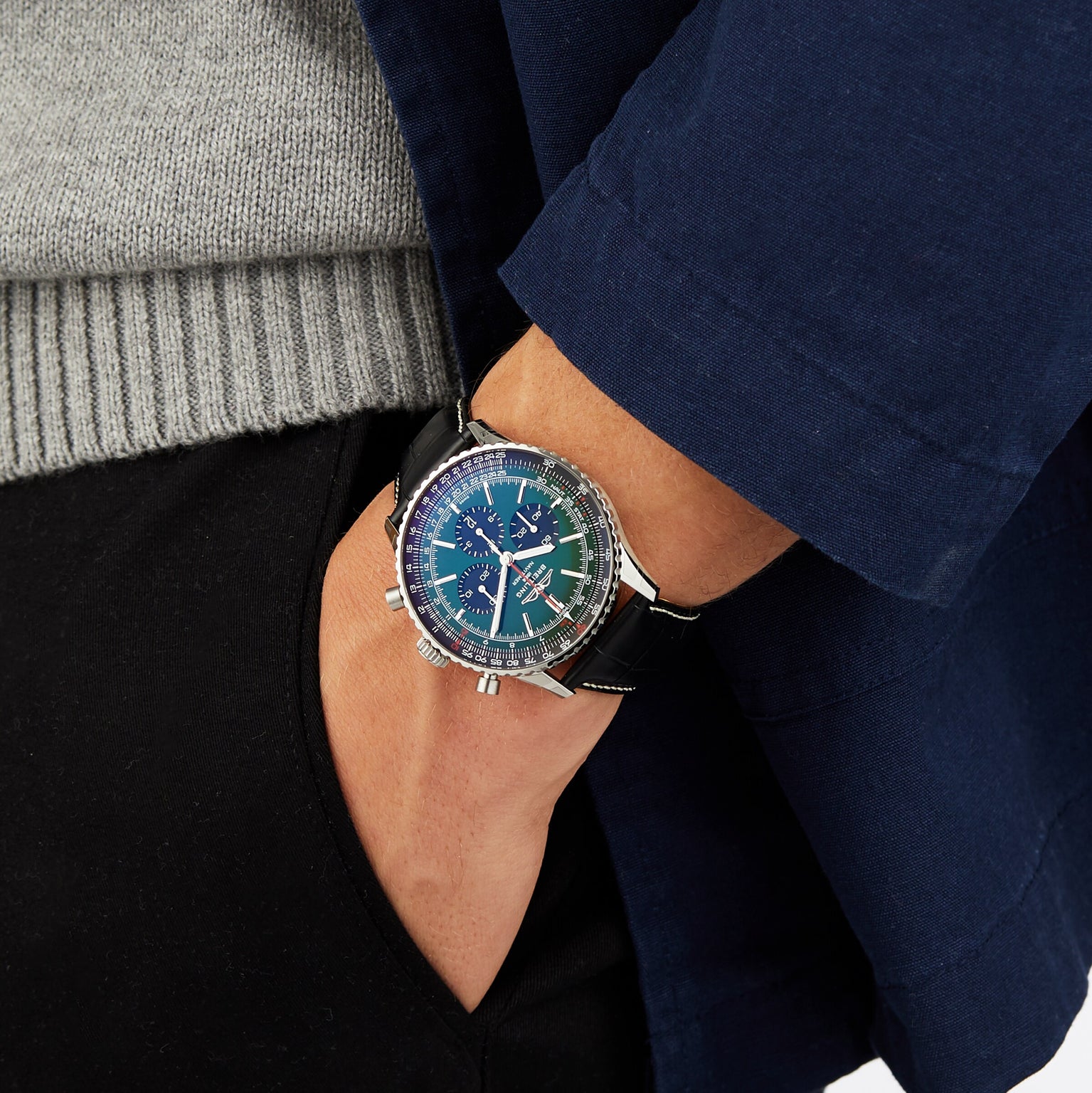 A person wearing a grey sweater and navy blue jacket sports a Breitling Navitimer B01 Chronograph 46, blue and silver dial, black band, with one hand in their pocket.