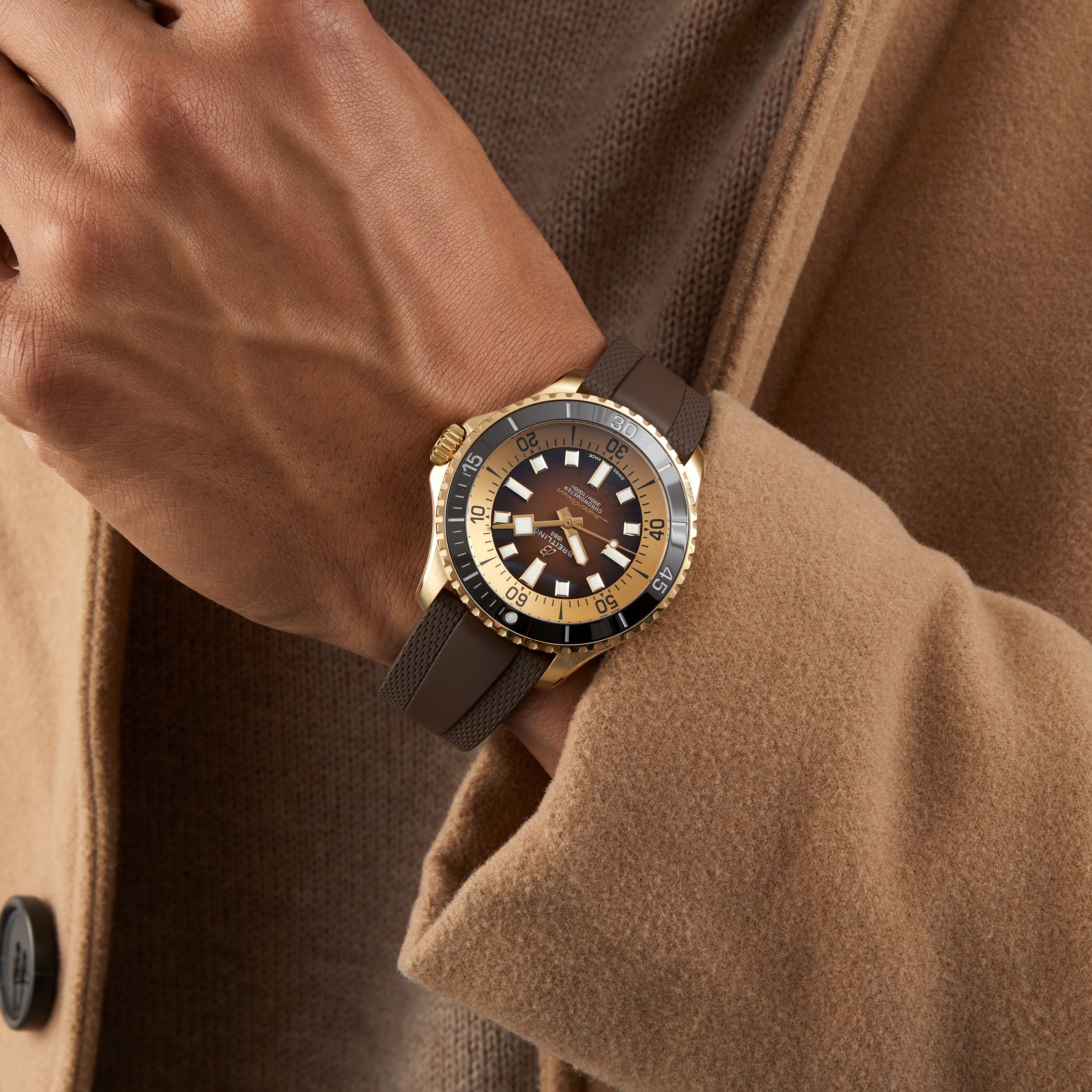 A person in a tan coat showcases a Breitling Superocean Automatic 44 wristwatch with a round face, bronze case, and textured brown strap on their left wrist.