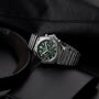 A Breitling Chronomat B01 42 The Watches Of Switzerland Group Centenary Exclusive wristwatch with a green and black dial rests on a dark surface beside a black wallet, headphones, and leather item. This limited edition features the famed B01 Manufacture Calibre.