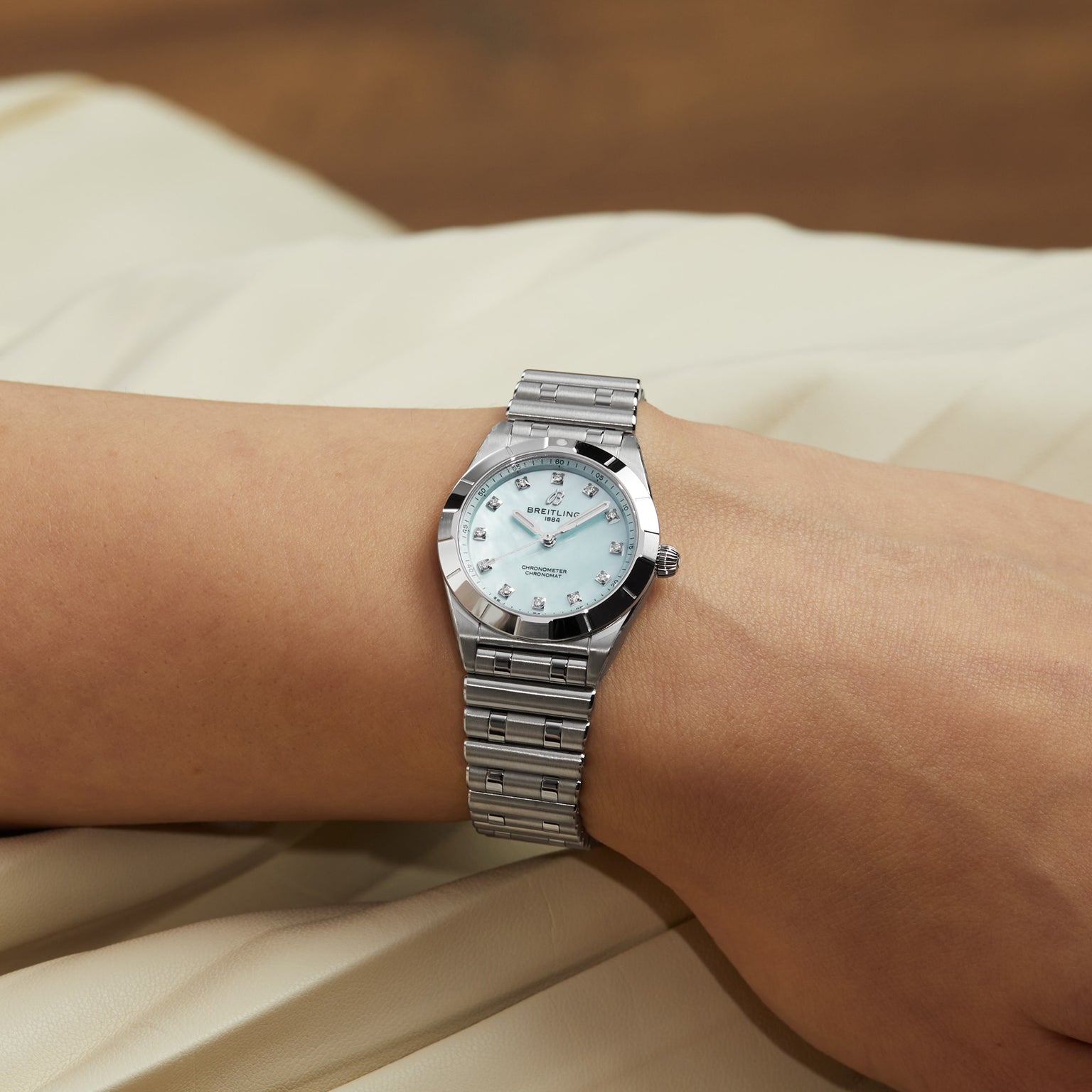 A Breitling Chronomat 28 wristwatch with a silver metal band and light dial is worn on a person’s wrist, resting on a pale fabric surface.